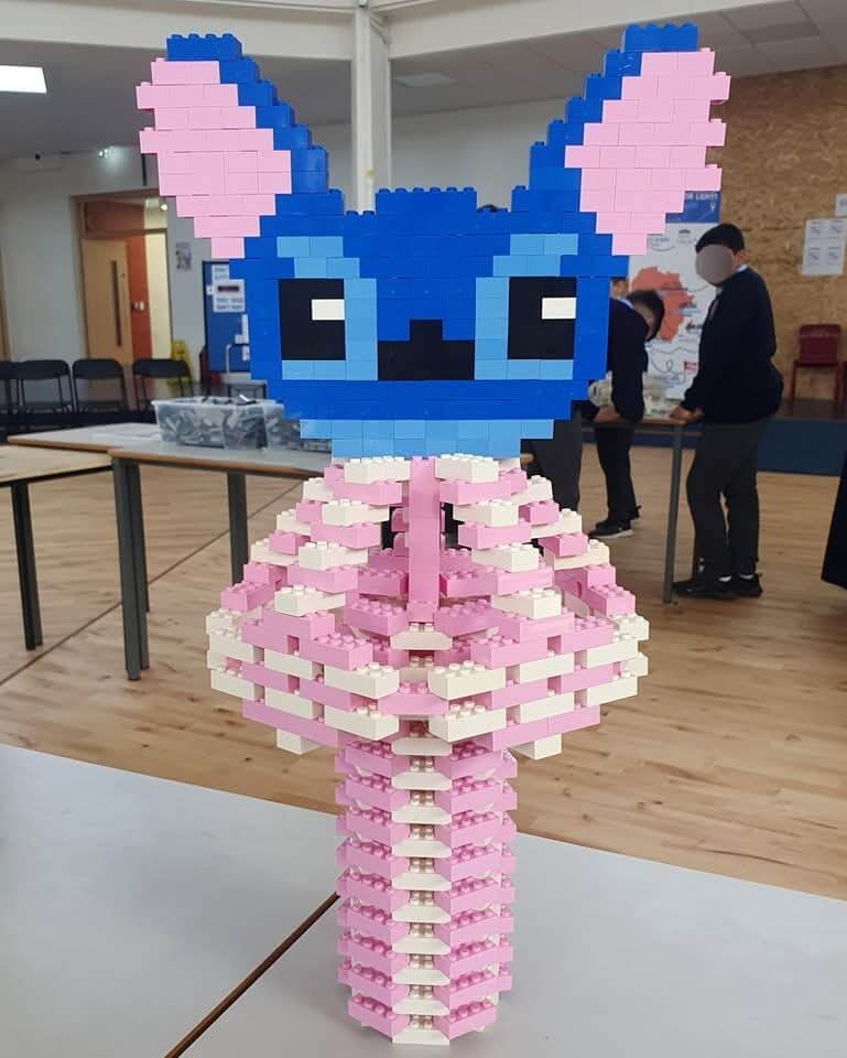 Large LEGO brick sculpture of a character with blue head and pink body displayed on a table in a hall.