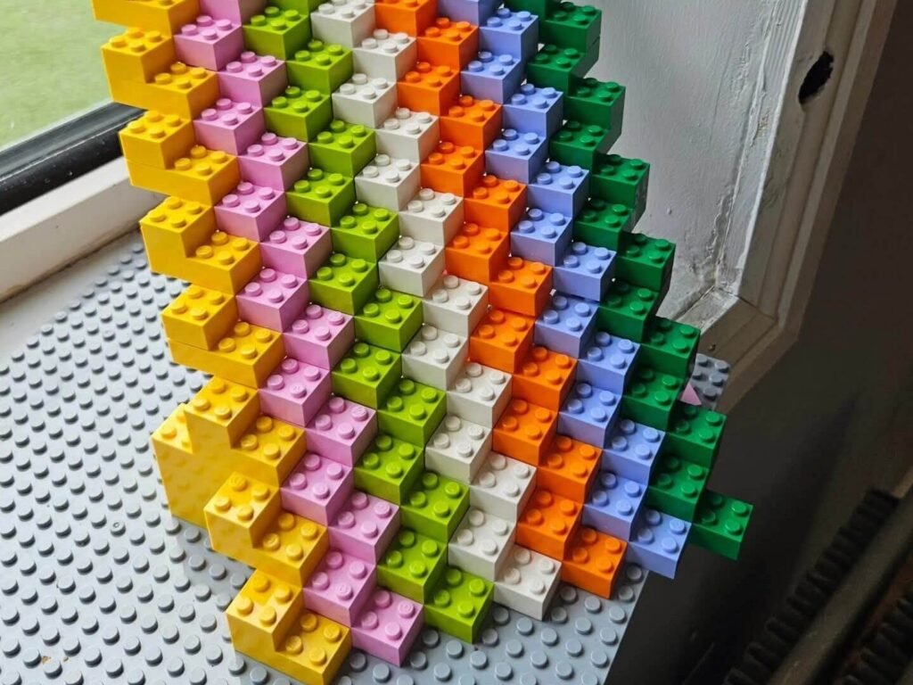 Stepped LEGO brick structure made from alternating coloured blocks placed on a baseplate beside a window.