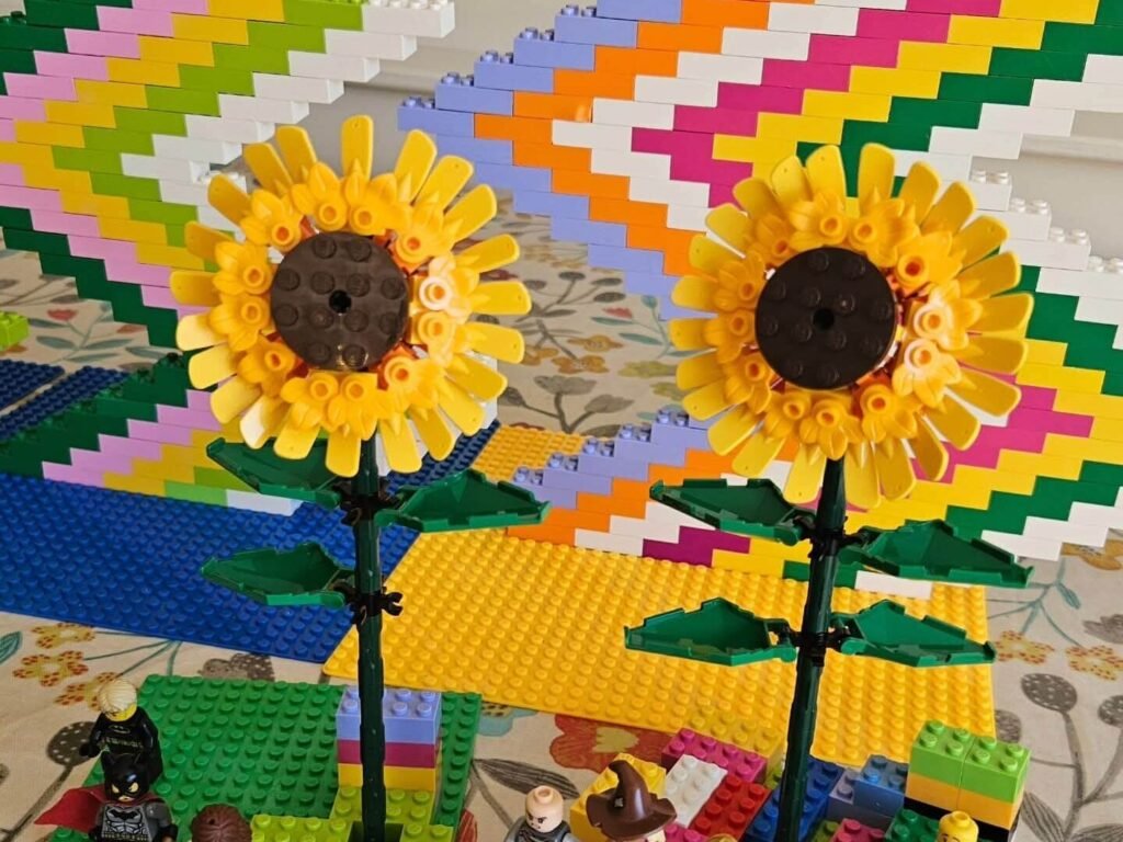 Two LEGO sunflower models built from yellow and green pieces, displayed in front of stepped multicolour brick panels.