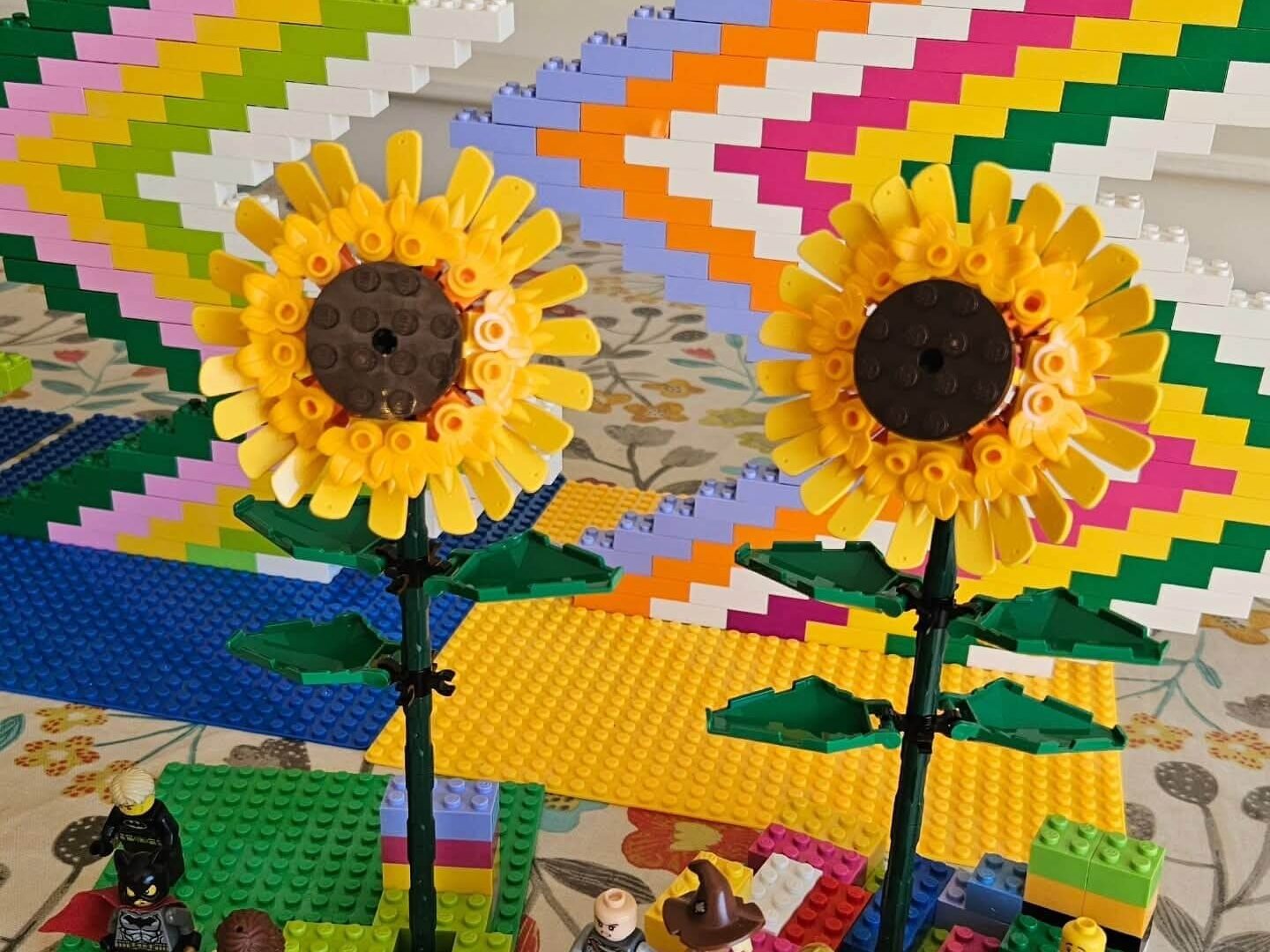 Two LEGO sunflower models built from yellow and green pieces, displayed in front of stepped multicolour brick panels.