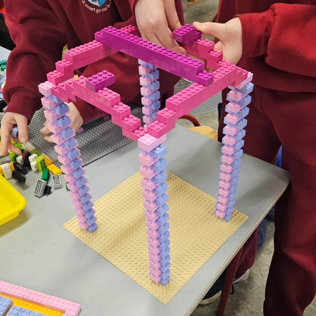 Children assembling a tall LEGO frame structure made from stacked pink and blue bricks on a baseplate.
