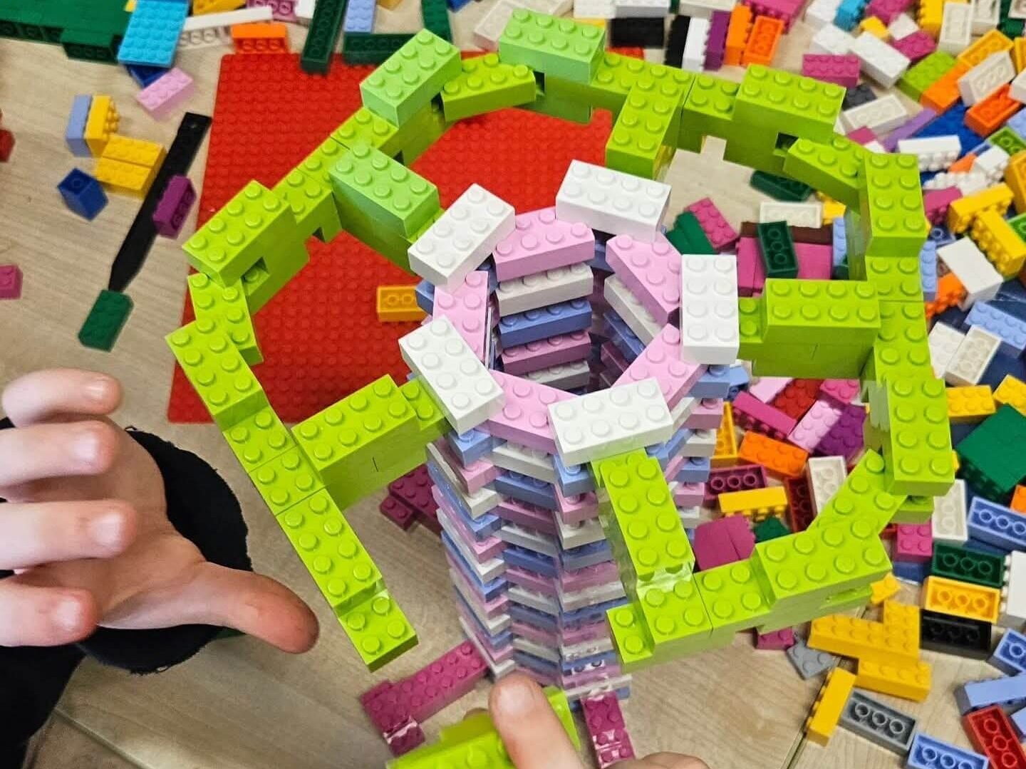 Hands assembling a circular LEGO structure with green outer elements and stacked inner bricks on a table.